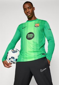 Maglietta da calcio verde a maniche lunghe con strisce orizzontali, con il logo del FC Barcelona e un logo Spotify nero. Pantaloni Nike neri.