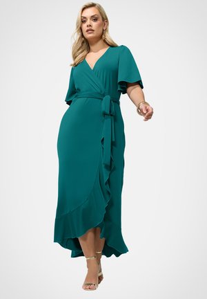 Kobieta ubrana w turkusową sukienkę kopertową z falbankami na rękawach i falbanami u dołu, zestawioną ze złotymi szpilkami z paskiem wokół kostki oraz złotą biżuterią.