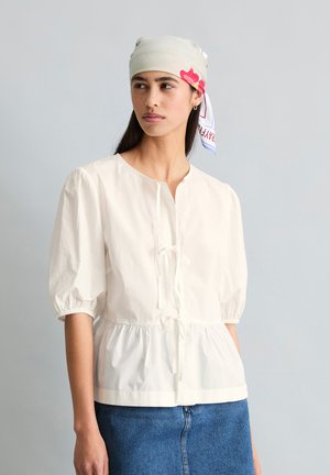 Kobieta ubrana w białą bluzkę z bufkami na rękawach i sznurkami, lekka chusta z czerwonym kwiatowym wzorem na głowie oraz niebieską spódnicę dżinsową.