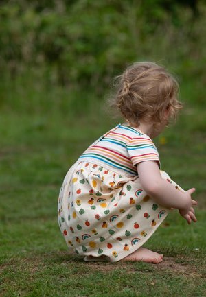 Toddler with braided hair squatting barefoot on grass, wearing a colorful striped shirt and a skirt with fruit and rainbow patterns.