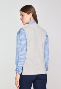 Gilet lavorato a maglia grigio chiaro con accenti blu a pois, indossato sopra una camicia a righe blu e bianca. Design con fessure laterali nella parte posteriore.