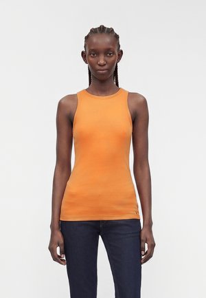Frau mit geflochtenem Haar, die ein ärmelloses orangefarbenes Top und dunkelblaue Jeans trägt, steht vor einem schlichten weißen Hintergrund.