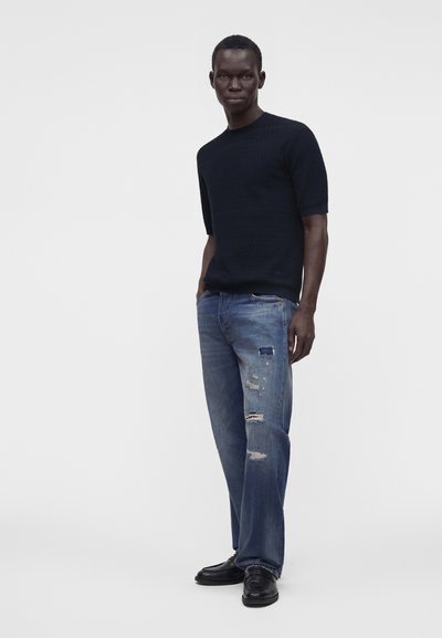 Homme portant un pull noir à manches courtes, un jean bleu déchiré et des chaussures noires, debout devant un fond clair uni.
