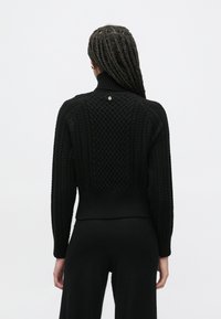 Crni pletivi džemper s visokim ovratnikom, s teksturiranim pletenim uzorkom i rebrastim rubom. Mali detalj logotipa na stražnjoj strani.