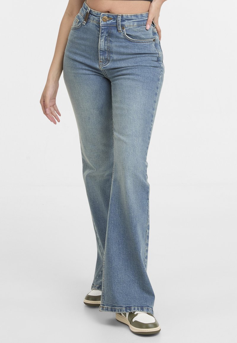 Femme portant un jean flare bleu clair taille haute avec fermeture à boutons et zip, associé à des baskets blanches et vertes.