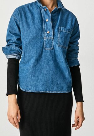 Persoon draagt een blauwe denim blouse met borstzak over een lange mouwen zwarte geribde jurk, de mouwen van het overhemd zijn opgerold.