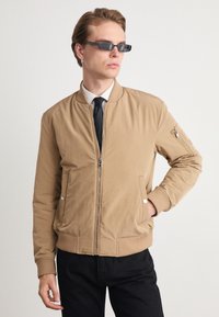 Veste bomber beige avec un zip sur le devant, des poignets et un ourlet côtelés, et des poches latérales. Elle est dotée d'une poche zippée sur le bras gauche. Portée avec une cravate noire.