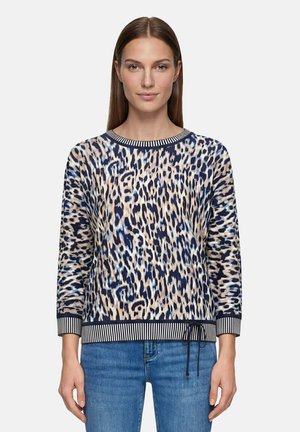 Femme portant un pull à manches longues imprimé léopard avec des poignets et un ourlet rayés, associé à un jean bleu, faisant face à l’avant sur un fond uni.