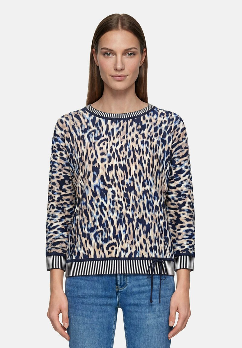 Femme portant un pull à manches longues imprimé léopard avec des poignets et un ourlet rayés, associé à un jean bleu, faisant face à l’avant sur un fond uni.