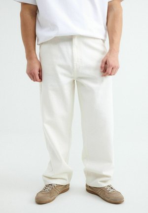 Mężczyzna w luźnych białych spodniach, beżowych trampkach i białej koszulce stojący na jednolitym białym tle.