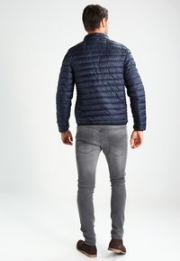 Marineblå puffjakke med kviltet design, tætsiddende silhuet og høj krave, parret med grå slim-fit jeans og brune ruskinds sko.