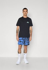 Črna bombažna majica s sloganom »Just Do It«. Modre kopalke z vzorcem batik. Bele nogavice in superge. Model stoji pred enobarvnim ozadjem.