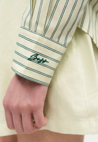 Poignet et main d'une personne portant un poignet de chemise rayé jaune clair brodé du texte vert « Brigitte ».