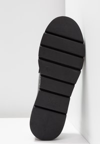 Semelle d'une chaussure noire avec de larges rainures horizontales et le nom de la marque "Xti" embossé près du talon.