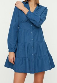 Donna che indossa un vestito lungo a maniche lunghe a portafoglio di colore blu, con una mano appoggiata sulla spalla opposta, su uno sfondo chiaro e semplice.