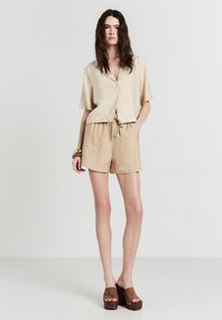 Donna con lunga chioma scura, vestita con una camicia corta beige con bottoni, pantaloni corti abbinati con lacci, sandali con piattaforma marroni e braccialetti, in piedi.