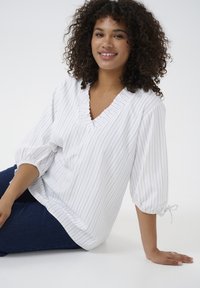 Weiße Bluse mit blauen vertikalen Streifen, V-Ausschnitt, Rüschensaum und 3/4 Puffärmeln mit elastischen Bündchen und Bändern. Glatte Textur.