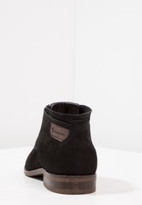 Bottine en daim noir avec un bout arrondi et un talon empilé de hauteur moyenne. Elle présente un accent en cuir marron près du talon.