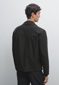 Black jacket with a smooth texture, featuring a collared neckline and a straight cut, viewed from the back. Minimalist design without embellishments.