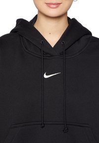 Svart hoodie med dragsko, prydd med en vit Nike-logotyp på bröstet. Materialet verkar mjukt med en slät textur.