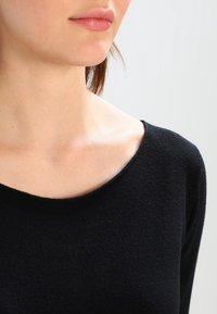 Schwarzer Pullover mit weicher Textur, ausgestattet mit einem weit ausgeschnittenen Bootsausschnitt und langen Ärmeln, die eng um den Ausschnitt und die Schultern anliegen.