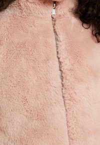 Nepbontjas in zachtroze, met een hoge kraag en een zilveren ritssluiting. Het oppervlak is gestructureerd en pluizig, met een gladde afwerking langs de ritssluiting.