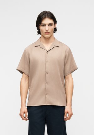 Νεαρός άνδρας με κυματιστά σκούρα μαλλιά φοράει μπεζ πουκάμισο με κοντά μανίκια και σκουρόχρωμο παντελόνι, στέκεται μπροστά σε απλό φωτεινό φόντο.