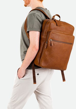 Bruine leren rugzak met een gestructureerde afwerking, ritszakken en verstelbare schouderbanden, gedragen op de rug van een persoon in casual kleding.
