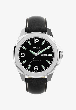 Ročna ura Timex z črnim usnjenim pasom, srebrno osemkotno ohišje, črn obraz, velike bele številke, svetleče kazalke in prikaz datuma.