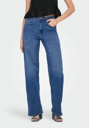 Person iført højtaljede vidde-ben blå jeans, sort flæset crop top, guldbøjle, og turkis åbne-tå blokhælede sandaler, hånd i lommen.