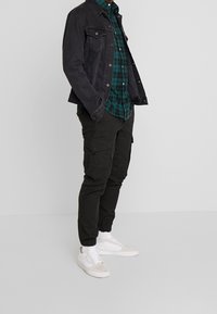 Zwarte denimjas, groene geruite overhemd met knopen, zwarte cargobroek en witte sneakers. Casual gelaagd met elastische manchetten op de broek.