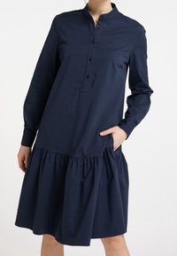 Abito blu navy a maniche lunghe con scollatura rotonda, bottoni frontali e gonna plissettata. Presenta tasche laterali e una superficie in cotone morbido.