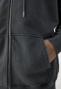 Felpa grigia con zip realizzata in tessuto testurizzato, dotata di tasca a marsupio, polsini a costine e una zip in metallo. Dettagli di cucitura sottili visibili.