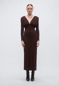 Καφέ μακρυμάνικο, εφαρμοστό φό�ρεμα με βαθύ V λαιμόκοψη και ρουά κορσέ. Λείος ύφασμα, μήκος μέχρι τον αστράγαλο, συνδυασμένο με μαύρες μπότες με τακούνι μέχρι το γόνατο.