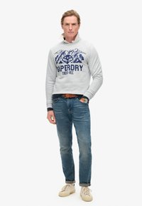 Felpa grigia con grafica di montagna navy e testo "Superdry". Abbinata a jeans blu e sneakers beige. Design casual, vestibilità comoda.