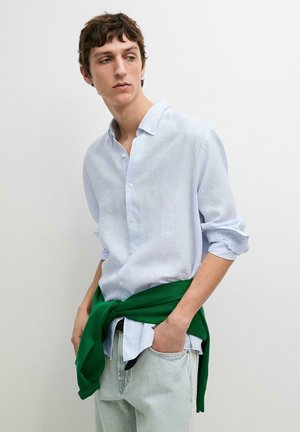 Ung mand iført lyseblå skjorte med opkrogede ærmer, grøn sweater bundet om livet og lyse jeans, stående op ad en hvid væg.