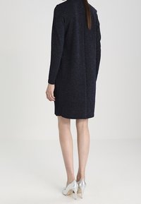 Navyblaues Kleid mit langen Ärmeln, strukturiertem Stoff, figurbetonter Silhouette und einer hinteren Naht. Kombiniert mit silbernen High-Heels.