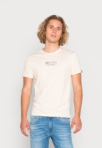 Beige bomulds t-shirt med korte ærmer, der har et lille mørkt logo i midten. Båret med lyseblå denim jeans.