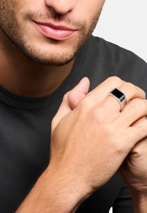 Homme avec une barbe légère portant une bague en argent avec une pierre noire rectangulaire à la main droite, portant une chemise sombre, mains jointes.