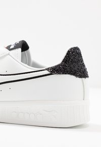 Bílé tenisky s černým třpytivým podpatkem, hladkým koženým svrškem, kontrastními černými pruhy a texturovanou gumovou podešví s logem značky.