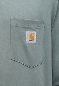 Ανοιχτό πράσινο μακρυμάνικο πουκάμισο με τσέπη στο στήθος και τετράγωνο λογότυπο Carhartt σε πορτοκαλί και άσπρα χρώματα. Ομαλή υφή.