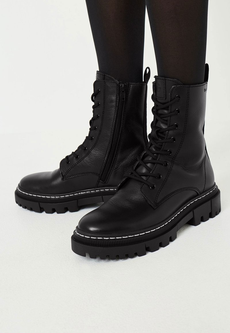 Bottes de combat noires en cuir à lacets avec fermetures éclair latérales et semelles à crampons épais, portées avec des collants noirs sur un fond clair.