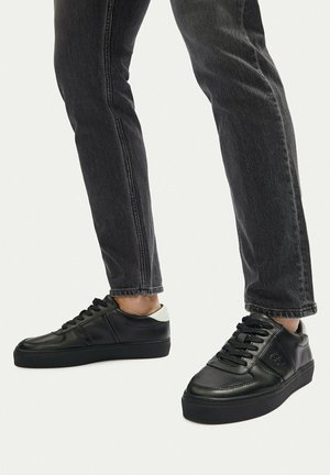 Persona con zapatillas de cuero negras con detalle blanco en el talón y vaqueros gris oscuro sobre un fondo claro y sencillo.