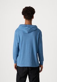 Felpa blu con una texture morbida, maniche lunghe e cappuccio con cordino. Presenta un orlo dritto e cuciture minime, indossata sopra pantaloni scuri.