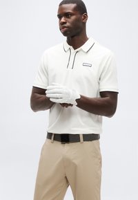 Άνδρας που φορά λευκό γάντι γκολφ, λευκό πουκάμισο πόλο με μαύρο περίγραμμα, μπεζ παντελόνι και σκούρα ζώνη, στέκεται μπροστά σε απλό φόντο.