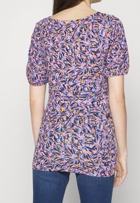 Fialový top s květinovým vzorem, vybavený nabíranými rukávy, kulatým výstřihem a přiléhavým pasem, vyrobený z lehké látky, kombinovaný s tmavými džíny.