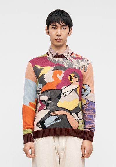 Πλεκτό πουλόβερ με πολύχρωμο αφηρημένο σχέδιο, σε αποχρώσεις πορτοκαλί, μαύρου και μπλε. Ριμπ μανσέτες και τελείωμα σε μπορντό χρώμα, φορεμένο πάνω από πουκάμισο με γιακά.