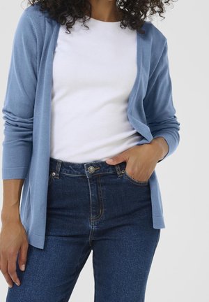 Frau trägt hellblauen Cardigan über weißem Hemd und dunkelblauen Jeans, mit einer Hand in der Tasche vor einfarbigem Hintergrund.