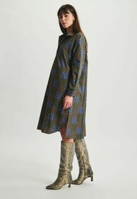 Robe à motifs multicolores avec des designs floraux bleus et verts sur un fond marron, dotée d'un col montant et d'une fente sur le côté.