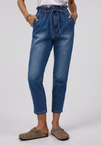 Blå denimbyxor med en samlad, dragkedja midjeresår och sidofickor; passar löst med uppvikta anklar; kombineras med bruna slip-on skor.